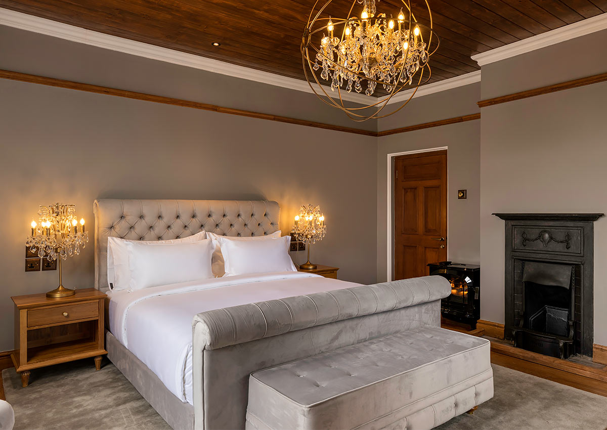 A Super Deluxe Room: a bed with a tufted grey headboard and footboard, crystal lamps on either side, a cast-iron fireplace and a chandelier under a timber ceiling